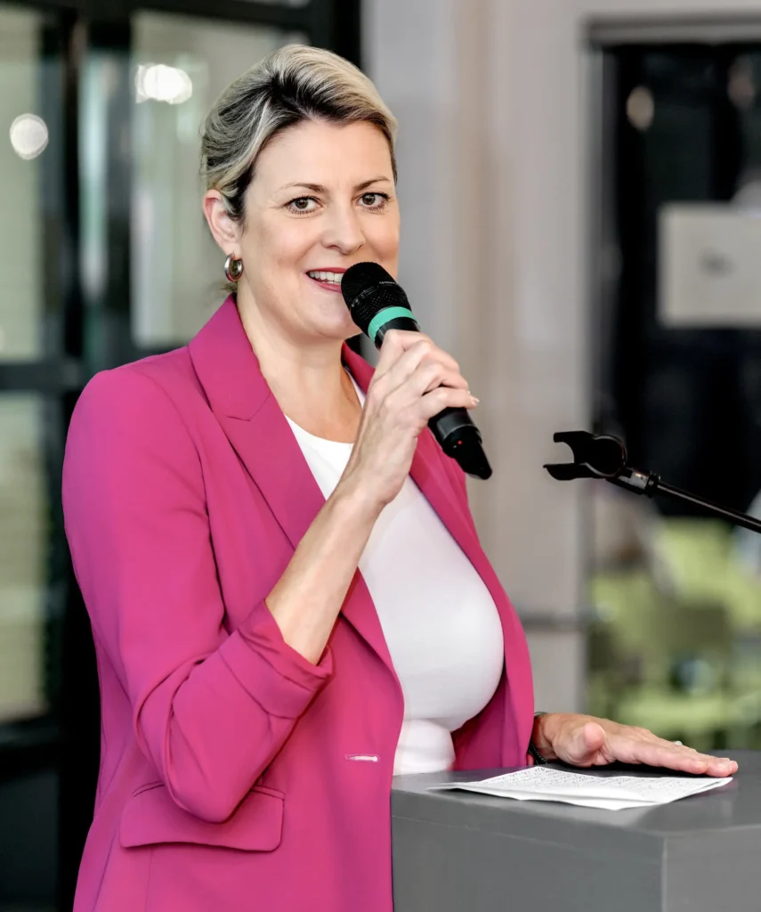 Rednerin im pinken Blazer spricht mit einem Mikrofon zu den Gästen